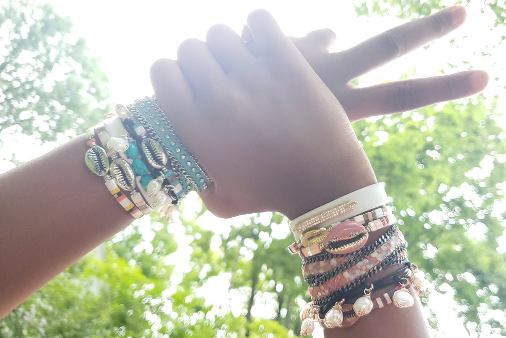 two hands holding each other , showing piece sign, both hands have multiple bracelets layered out . one hand has blush pinkand white bracelets and other hand has turquoise color bracelets