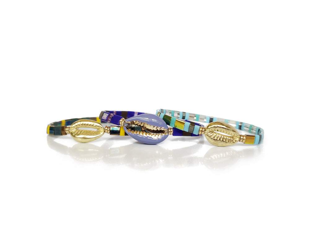3 coerie shell bead stretch bracelets.Top bracelet in light blue beads and accented gold cowrie shell bead.Middle bracelet is in dark blue beads and has  gold and blue enameled cowrie shell bead .Bottom bracelet is in dark blue green beads and has gold cowrie shell bead accent.