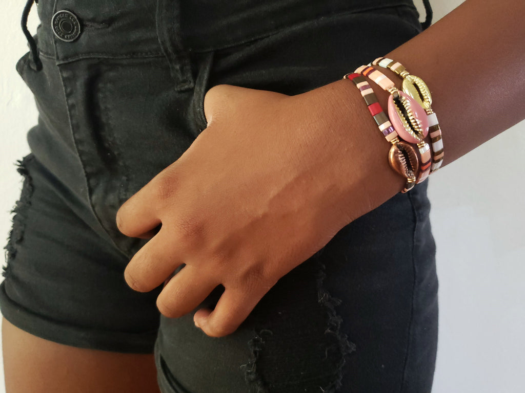 Girl is wearing blackshorts, hers hand is in a pocket and she has pinkblushbracelet set of 3 , bracelets have cowrie shells in gold , copper and enameled pink.