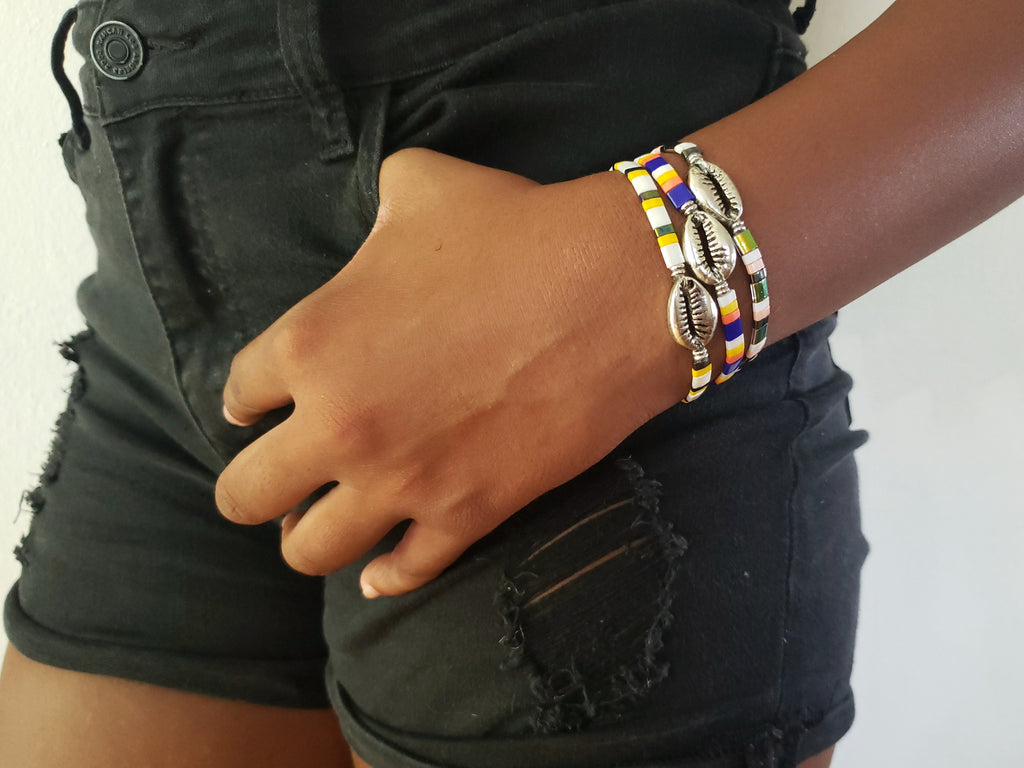 Girl is wearing black shorts, hers hand is in the pocket and she has 3 silver color cowrie shell stretch bracelets with rectangle beads in white/blue/pink and yellow.